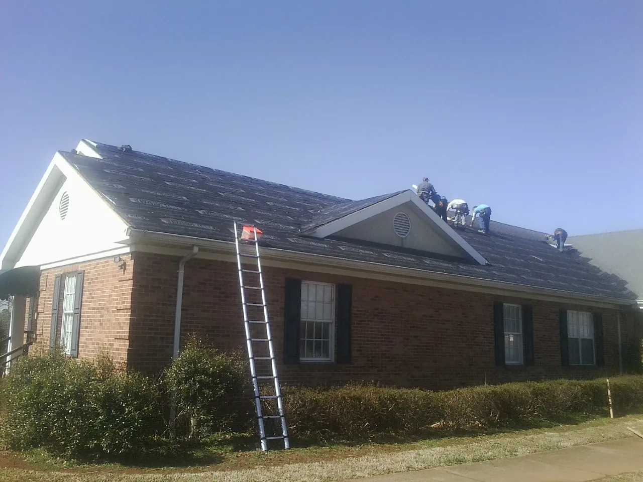 Crew tearing shingles off a brick ranch roof with a ladder set — Emergency Roof Repair work by our crew in Ehrhardt
